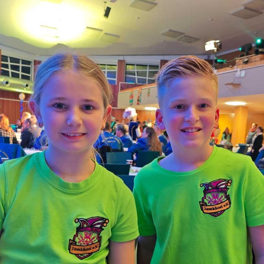 Zwei Kinder in grünen Vereins-T-Shirts mit Narrenkappe sitzen lächelnd in einem bunt beleuchteten Veranstaltungsraum.