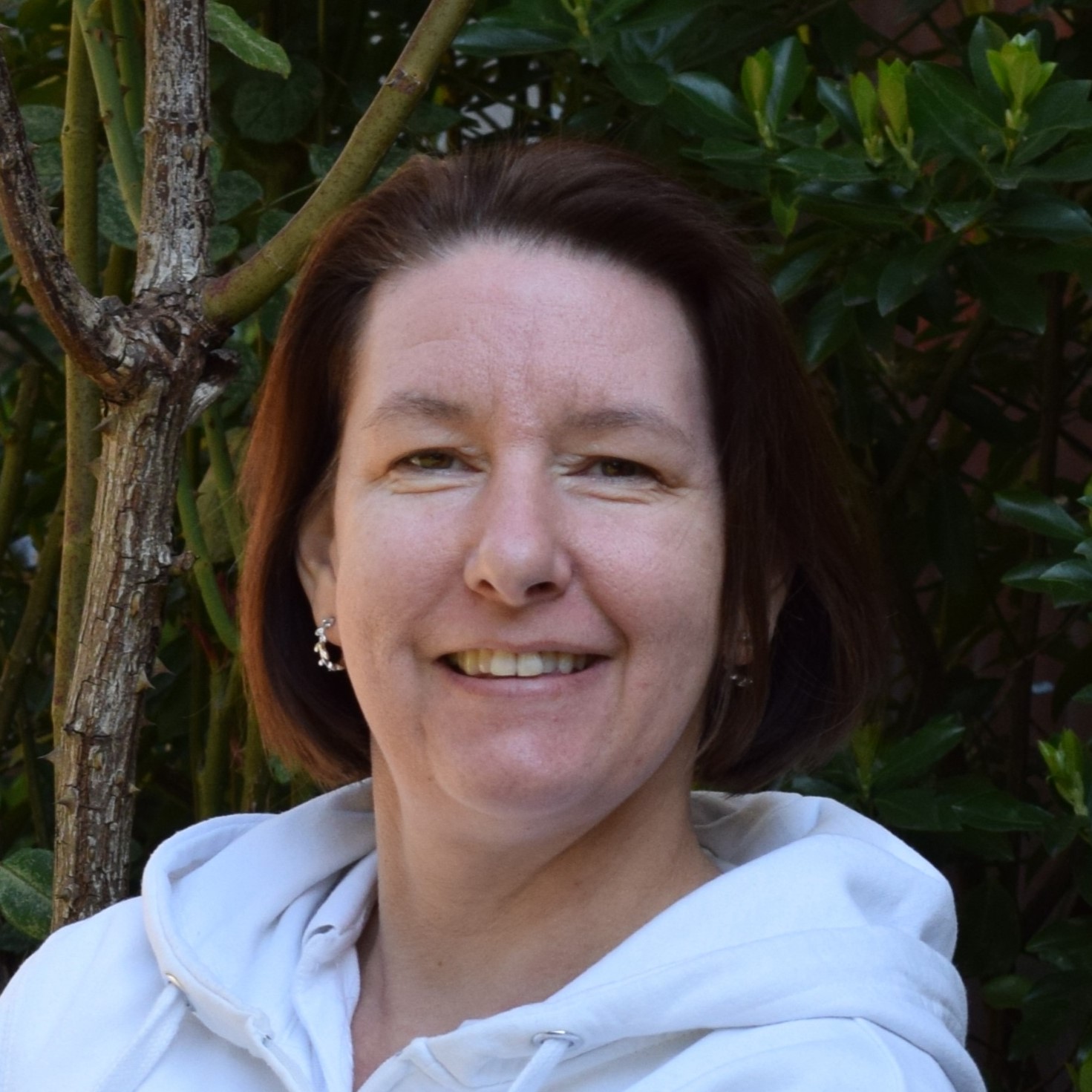 Frau mit kurzem braunem Haar, weißem Hoodie und silbernen Stern-Ohrringen lächelt freundlich vor grünem Busch.