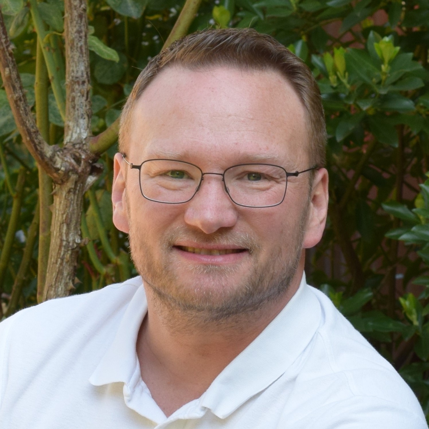 Mann mit kurzem Haar, Brille und weißem Polohemd lächelt leicht zur Kamera, im Hintergrund grünes Blattwerk und Äste.