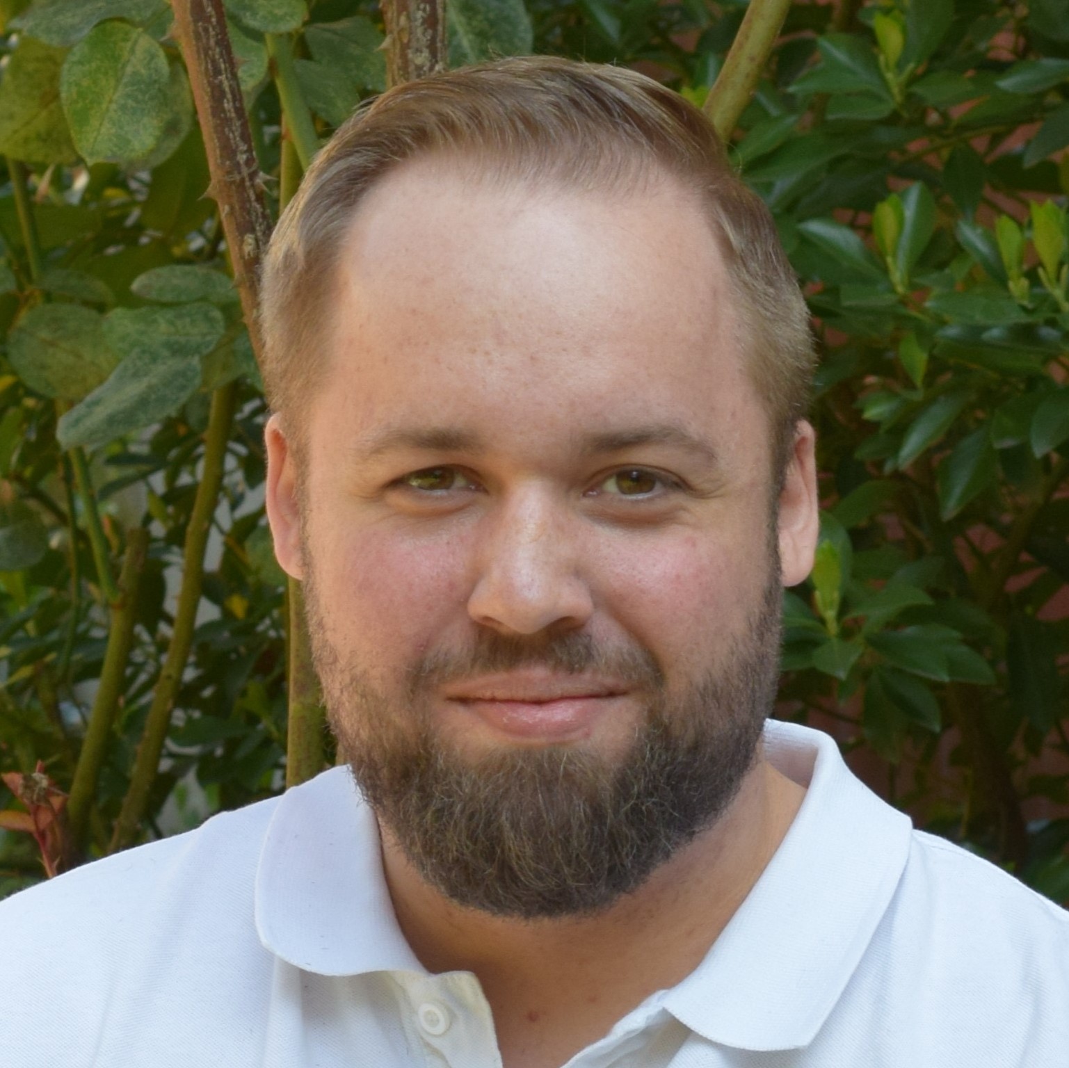 Mann mit blondem Haar, kurzem Bart und weißem Polohemd blickt ernst in die Kamera, grünes Blattwerk im Hintergrund.