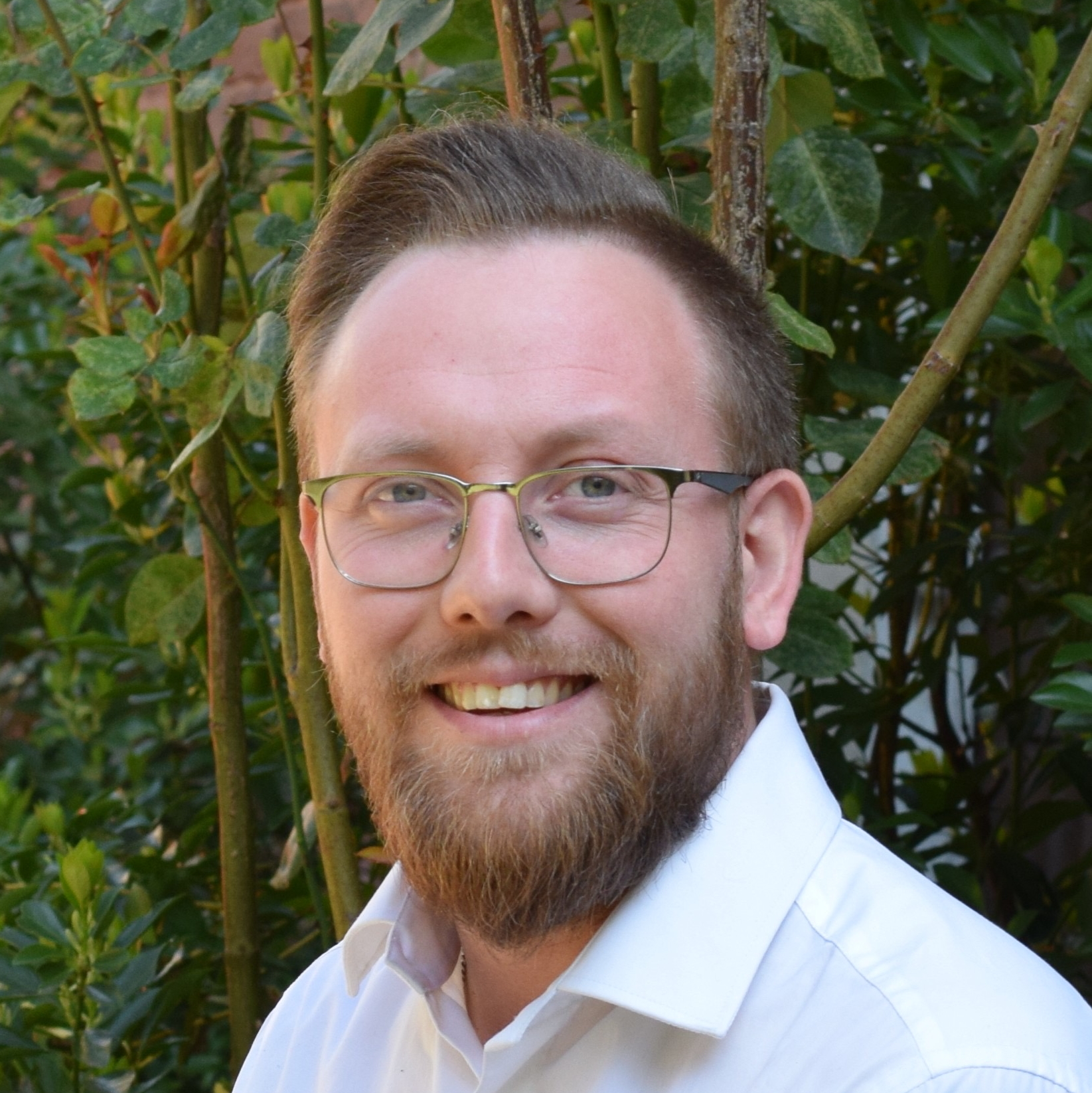 Porträt eines freundlich lächelnden Mannes mit Brille und Bart vor grünem Gartenhintergrund.