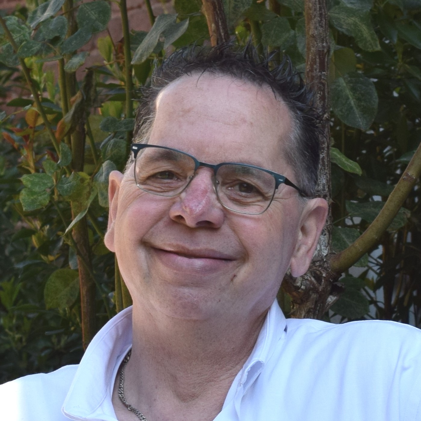 Mann mit Brille und kurzem dunklem Haar lächelt herzlich in weißem Hemd vor grüner Hecke.