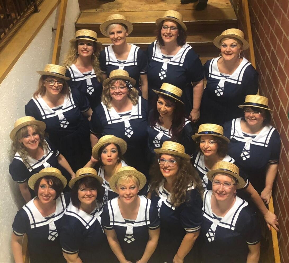 Gruppe von 15 Damen in Matrosenkostümen mit Strohhüten steht lächelnd auf einer Treppe und blickt zur Kamera.