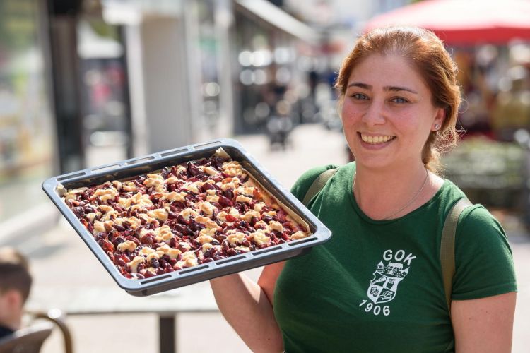 Eine Frau im grünen T-Shirt mit GOK-Wappen hält ein Blech Kirschkuchen, lächelt und steht in einer Fußgängerzone.
