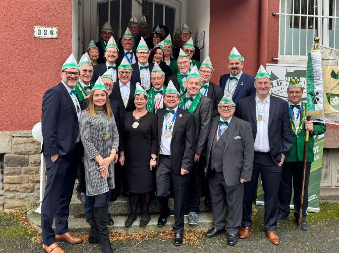 Große Gruppe in Anzügen und grünen Karnevalshüten steht vor Haus und lächelt zur Kamera.