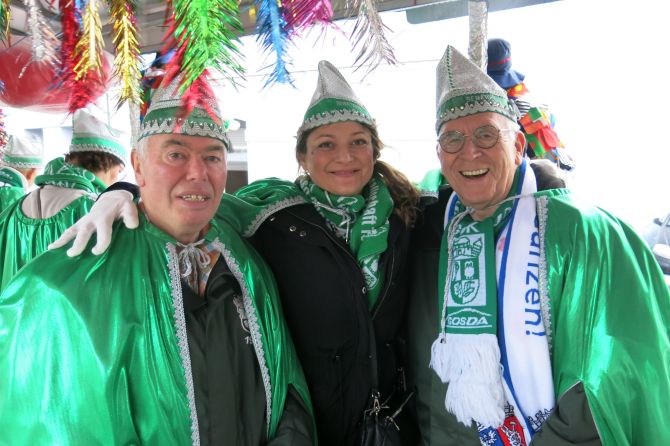 Drei GOK-Mitglieder in grünen Umhängen und Karnevalshüten feiern gemeinsam auf einem bunt geschmückten Karnevalswagen.