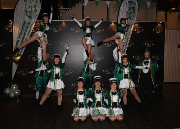 Tanzgruppe in grünen Kostümen bildet akrobatische Pyramiden vor schwarzer Bühne mit „Atrium“-Logo.